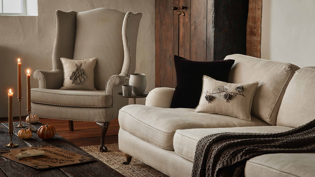 Create a warm Halloween display with a beige sofa, striped armchair, lit candles, blanket, wooden coffee table with small pumpkins and ouija board, plus the Coral & Tusk Bat Branch Pillow (ships in one week) for added charm.