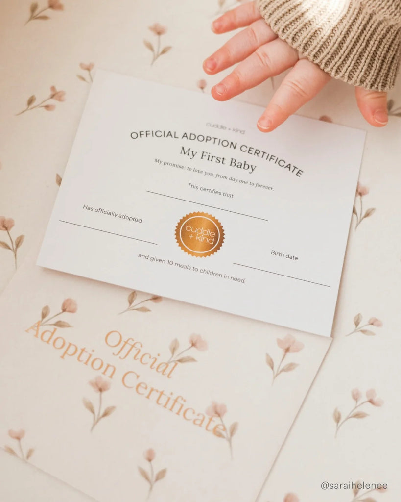 A baby’s hand touches an "Official Adoption Certificate" beside the Cuddle + Kind My First Baby (Bluebell) doll on a floral surface. The certificate displays a gold seal and love promise. @sarahhelenee is visible in the image. Ships in one week.