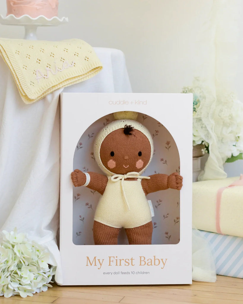 Cuddle + Kind My First Baby (Buttercup), a fair trade, hand-knit brown-skinned doll in a cream outfit and bonnet, is made from natural cotton yarn and helps feed 10 children with every purchase. Ships in one week.