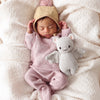 A sleeping baby, dressed in a cozy pink knit outfit and a beige hood with bear ears, lies nestled in a soft, off-white blanket. The baby is holding the Cuddle + Kind Baby Limited Edition Grey Bat made from Peruvian cotton yarn, adding a touch of whimsy to the serene scene. These hand-knit baby animals are truly collectible treasures.