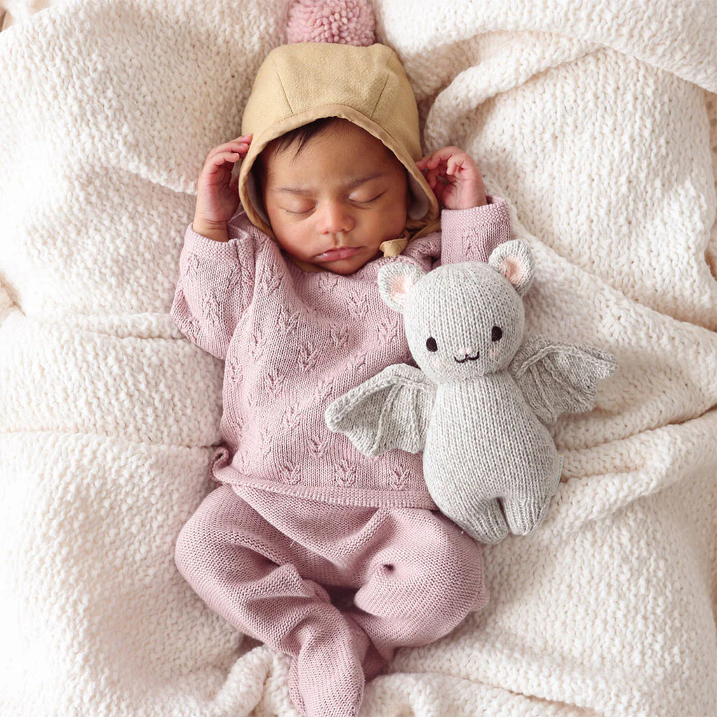 A sleeping baby, dressed in a cozy pink knit outfit and a beige hood with bear ears, lies nestled in a soft, off-white blanket. The baby is holding the Cuddle + Kind Baby Limited Edition Grey Bat made from Peruvian cotton yarn, adding a touch of whimsy to the serene scene. These hand-knit baby animals are truly collectible treasures.