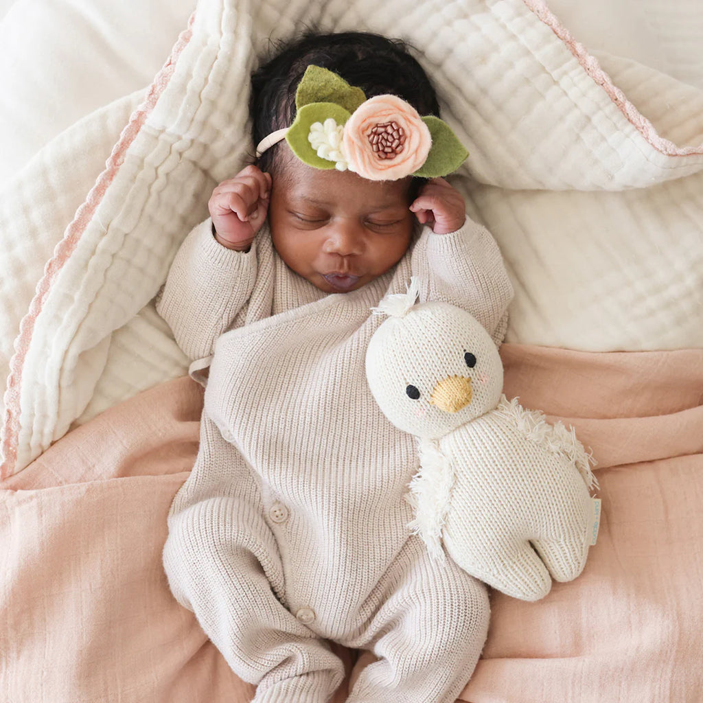 A newborn baby sleeping peacefully, swaddled in a pink blanket with a Cuddle + Kind Baby Gosling beside her. She wears a cute headband with a large floral decoration.