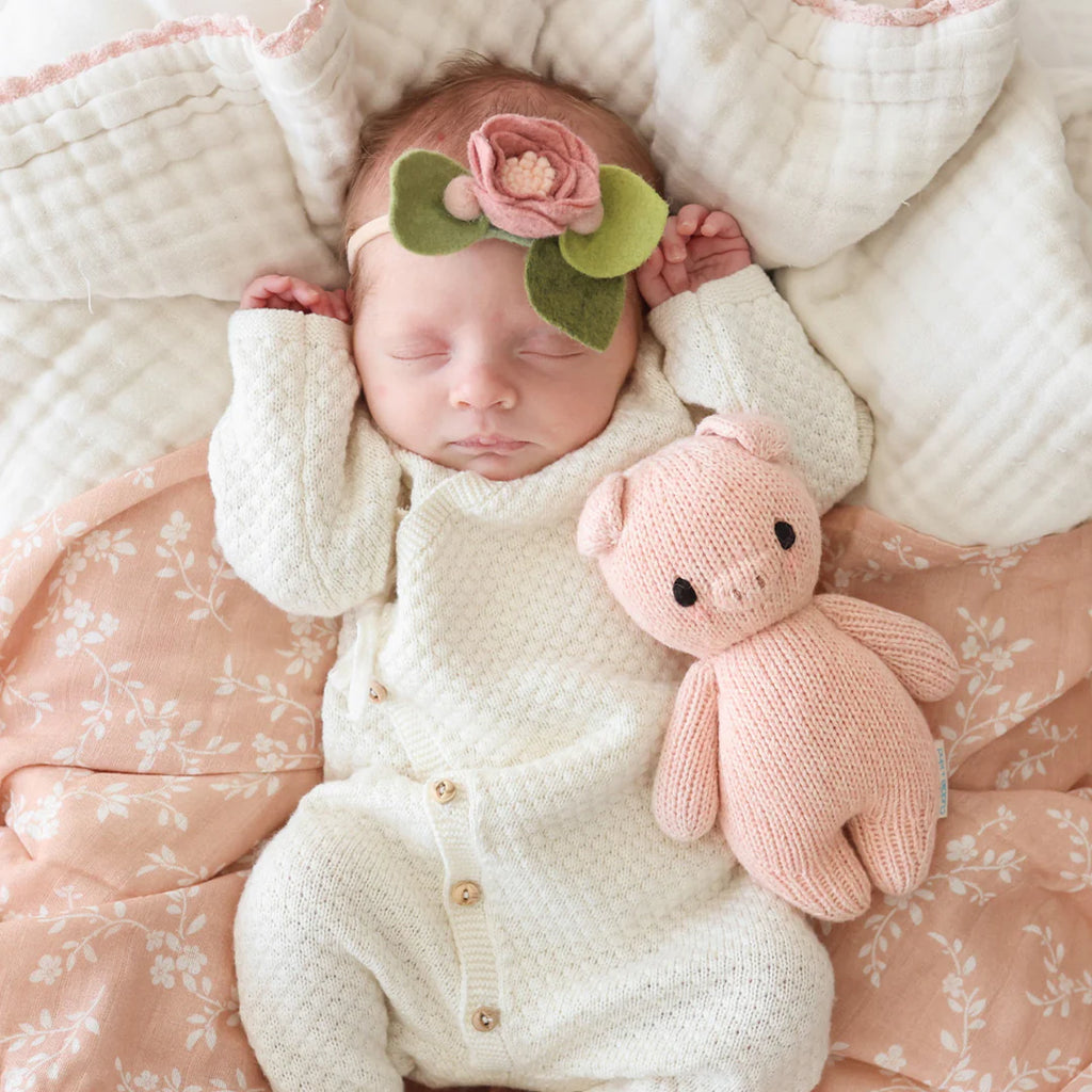 Newborn baby sleeping in a cozy pink blanket, wearing a hand-knit white outfit and a headband with a large flower, holding a Cuddle + Kind Baby Piglet toy.