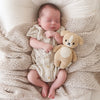 A newborn baby sleeps peacefully on a knitted blanket, clutching a small Cuddle + Kind Baby Puppy hand-knit from Peruvian cotton yarn, dressed in a beige knitted romper.