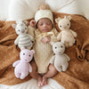 A newborn baby sleeps peacefully on a bed surrounded by plush animal toys, including a Cuddle + Kind Baby Zebra, giraffe, elephant, and a rabbit hand-knit from Peruvian cotton yarn. The baby is wearing
