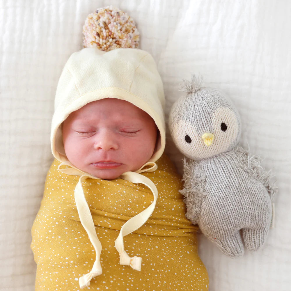 Newborn baby swaddled in a yellow blanket and wearing a cream hat with a pom-pom, lying next to a Cuddle + Kind Baby Penguin on a white background.