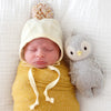 Newborn baby swaddled in a yellow blanket and wearing a cream hat with a pom-pom, lying next to a Cuddle + Kind Baby Penguin on a white background.