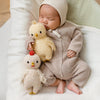 A sleeping baby in a beige knit outfit cuddles two heirloom-quality Cuddle + Kind Baby Chicken dolls—one yellow, one white—while lying on a soft white blanket.