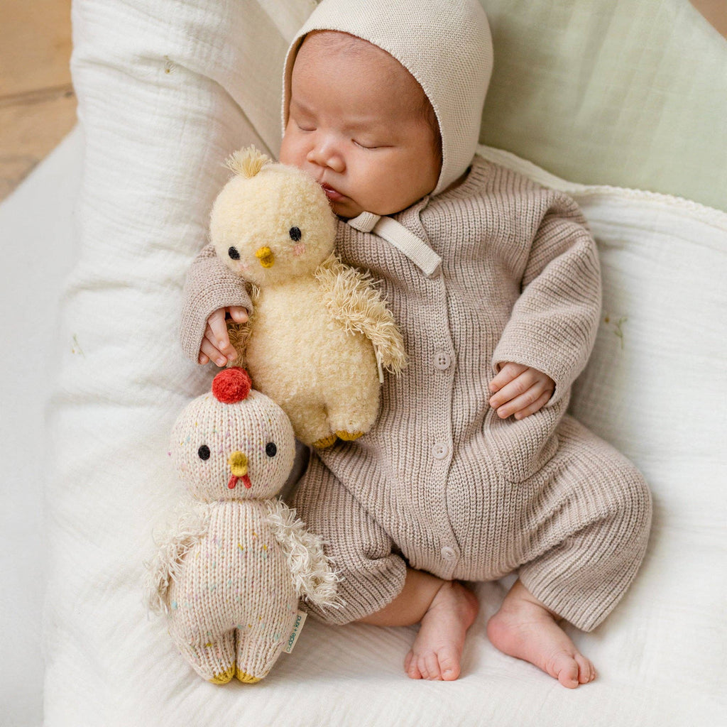 A sleeping baby in a beige knit outfit cuddles two heirloom-quality Cuddle + Kind Baby Chicken dolls—one yellow, one white—while lying on a soft white blanket.