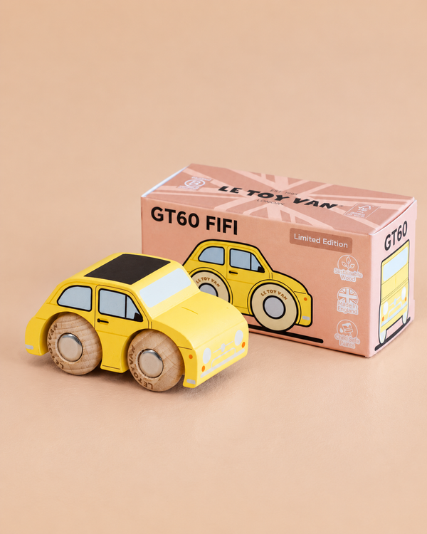 A classic miniature yellow wooden toy car with black windows sits beside its pink and brown packaging box labeled “No. 10 GT60 Fifi | Made in England” on a beige background. The box features charming illustrations of the vintage toy car.