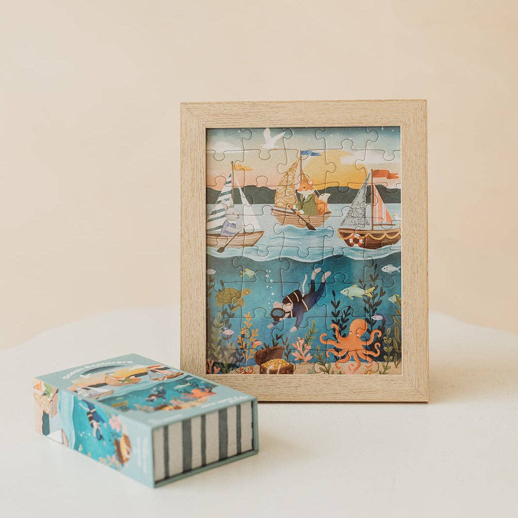 The Ocean Explorers 40 Piece Mini puzzle is shown completed and framed, featuring sailboats gliding on the water as well as a diver, colorful fish, and an octopus beneath the waves. Its matching puzzle box sits beside the frame on a white surface against a beige background—perfect for providing kids with hours of creative jigsaw fun.