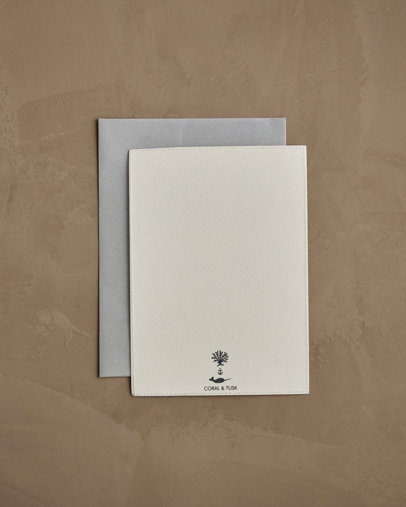 The Coral & Tusk Peek a Tree Card, featuring "CORAL & TUSK" and a small Christmas tree logo on white textured cardstock, rests atop a gray envelope on a light brown textured surface.