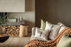 A cozy living room features a sofa adorned with festive pillows, including the Coral & Tusk Christmas Tree Car Pocket Pillow, a chunky brown knit blanket, green cushions, woodland animal decor, decorative trees, firewood, and a wooden stool.