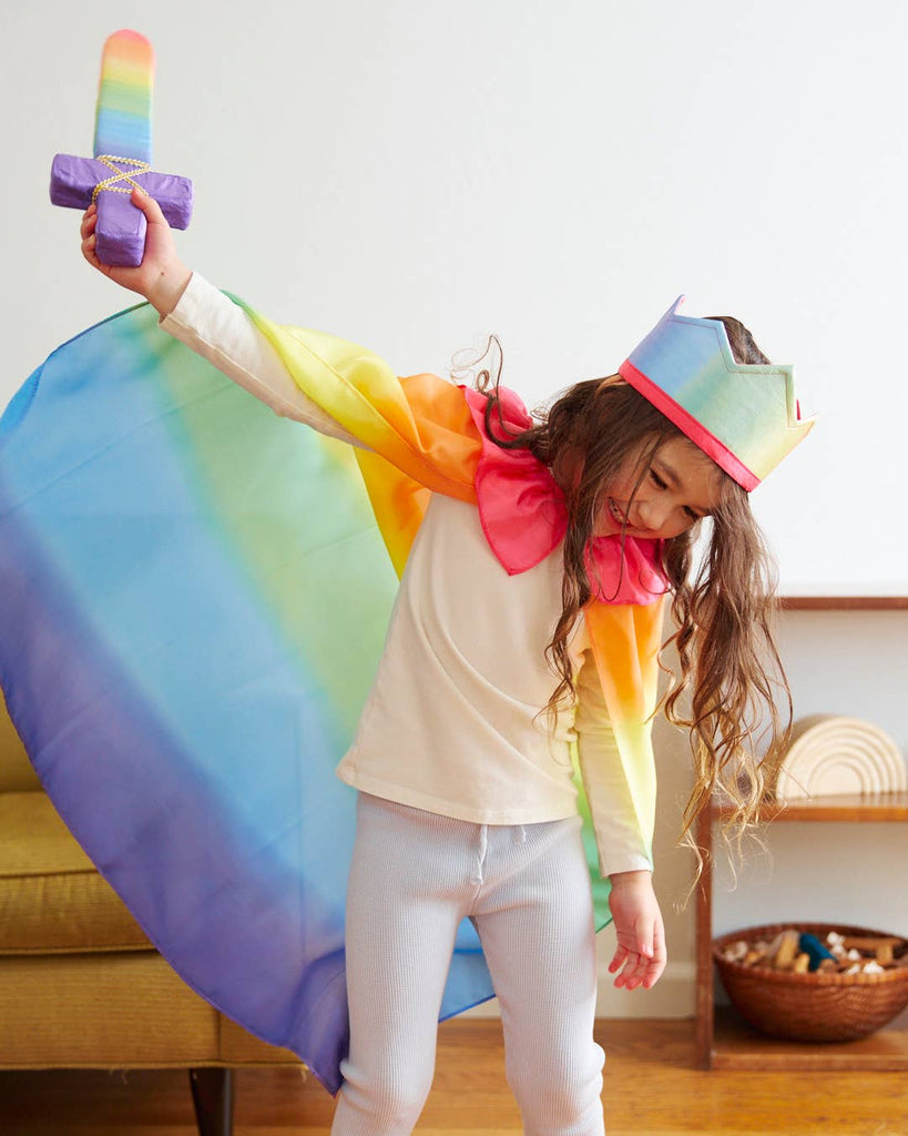 A child, draped in a colorful cape and crown, happily immerses themselves in pretend play with Sarah's Silk Soft Sword, beautifully decorated with rainbow designs. This lively scene takes place in a room featuring a yellow armchair and wooden shelves in the background, where imagination truly knows no bounds.