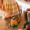 A child in a yellow plaid dress reaches for sliced avocado and papaya arranged in small bowls on the Cathy the Carrot Plate & Spoon Set, made from Hevea wood, beside spice jars and a cucumber, enjoying mealtime with this non-toxic baby tableware.