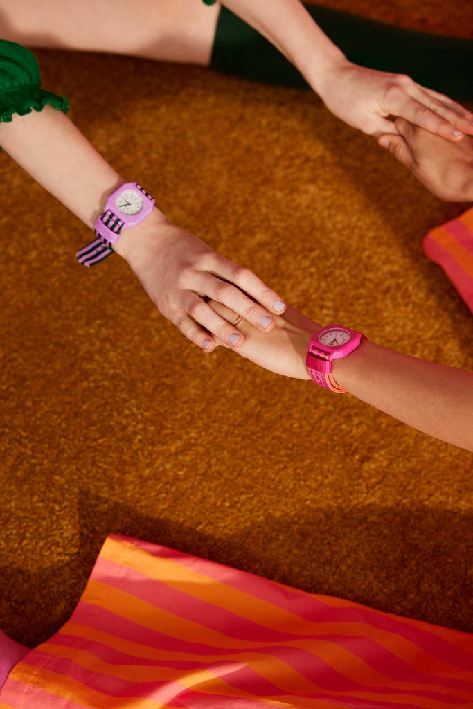 Two people sit on a brown carpet, holding hands. Each wears a colorful Mini Kyomo Watch - Lavender Field with Japanese movement—one purple striped, one solid pink. Bright skirts and green socks peek out below.