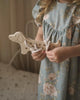 A young child in a light blue floral dress holds a **Maple Wood Lacing Toy - Rex the Dog** made from Maple wood with white cotton laces threaded through its holes. The scene is softly lit with warm tones, and the background shows a bed with a white polka-dotted bedspread.