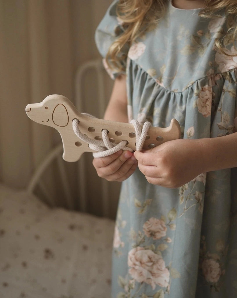 A young child in a light blue floral dress holds a **Maple Wood Lacing Toy - Rex the Dog** made from Maple wood with white cotton laces threaded through its holes. The scene is softly lit with warm tones, and the background shows a bed with a white polka-dotted bedspread.