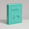 A turquoise, book-shaped box titled "Board Game: Catch Phrase Vintage Bookshelf Edition" stands upright on a white surface, featuring two cartoon characters interacting on the front.