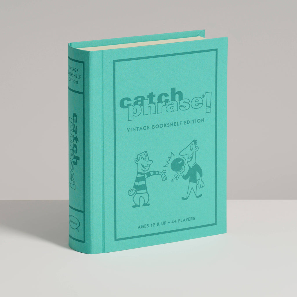 A turquoise, book-shaped box titled "Board Game: Catch Phrase Vintage Bookshelf Edition" stands upright on a white surface, featuring two cartoon characters interacting on the front.