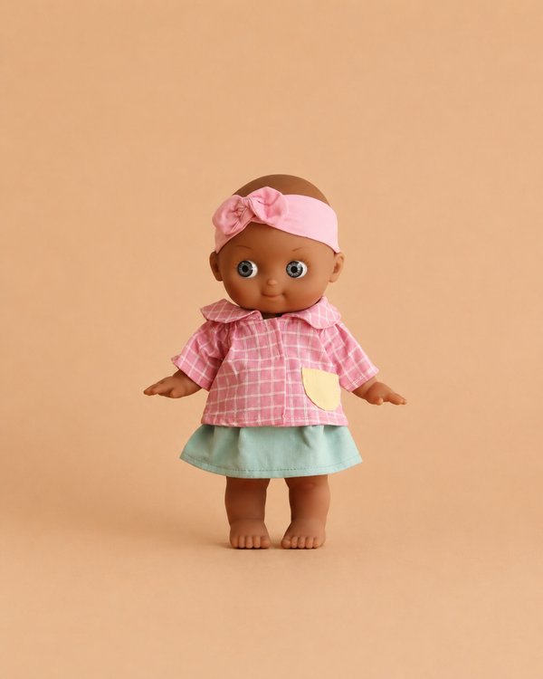 The Minikane Mini Pancha Doll - Maia with Pink Checkered Outfit features a high-quality vinyl design with brown skin, dressed in a pink checkered shirt, pale green skirt, and a pink headband with bow, standing barefoot against a beige background—perfect for inspiring imaginative play.