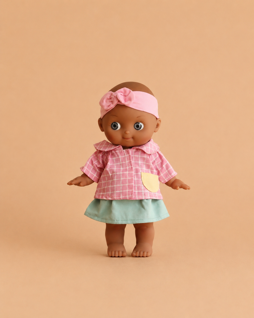 The Minikane Mini Pancha Doll - Maia with Pink Checkered Outfit features a high-quality vinyl design with brown skin, dressed in a pink checkered shirt, pale green skirt, and a pink headband with bow, standing barefoot against a beige background—perfect for inspiring imaginative play.
