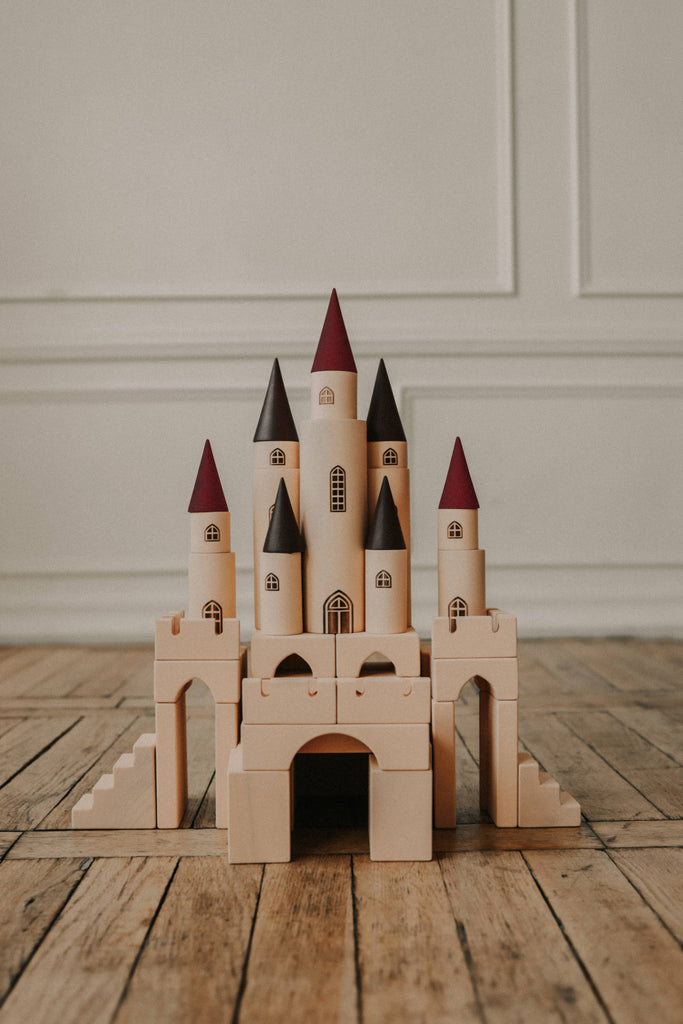 The Great Kingdom Building Set whimsically adorns a smooth floor with its tan blocks and towers, topped by dark and light red pointed roofs. Each piece is crafted with smooth edges and coated in non-toxic paint, set against a white paneled wall.