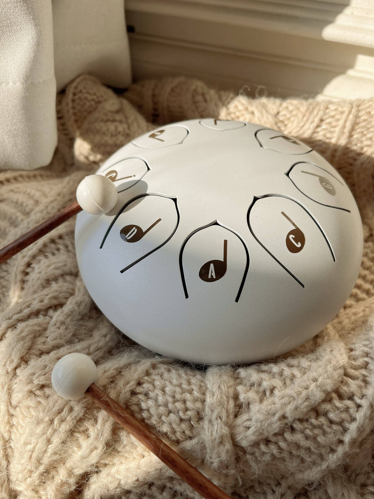 A Steel Tongue Drum in white, labeled with musical notes, sits on a knitted beige blanket beside two wooden mallets—an elegant musical toy instrument in soft sunlight.