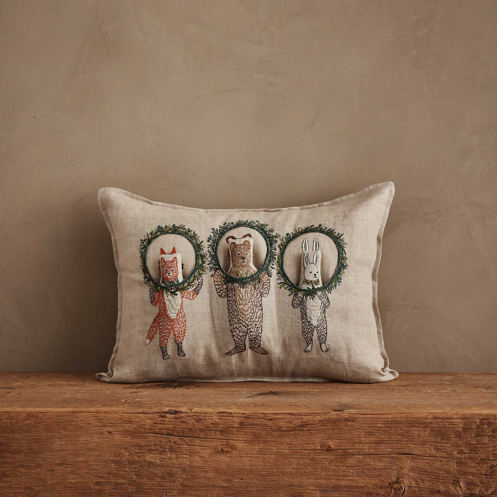 The Christmas Wreath Trio Pocket Pillow is a decorative beige pillow with embroidered woodland friends—a fox, bear, and rabbit—holding green wreaths, perfect for adding holiday charm to your rustic decor.
