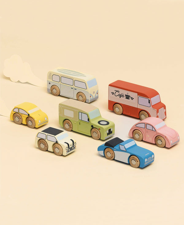 Seven Vintage Toy Cars, including a café van, are arranged on a beige background. Crafted from FSC-certified wood, they evoke classic automobile charm. A white cloud cut-out behind the yellow car suggests motion.