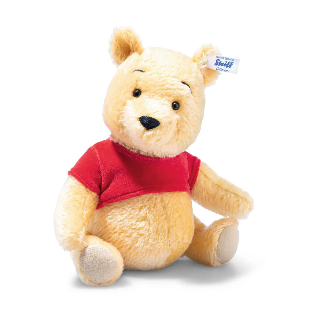 A Steiff plush toy of Disney's Winnie the Pooh sitting, with a red shirt and a small tag on its ear, displayed against a white background.