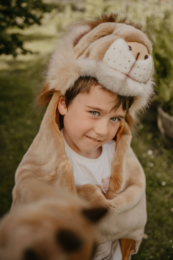 A young boy outdoors wears the Costume - Lion by Wild & Soft—a plush lion hood and cape—smiling gently with one arm outstretched as if snapping a selfie, set against a grassy background with greenery and blurred trees.