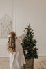 A young girl with long brown hair in a light dress decorates a small Christmas tree with Sabo Concept Christmas Ornaments and lights in a cozy, softly lit room with light walls and wooden floors.