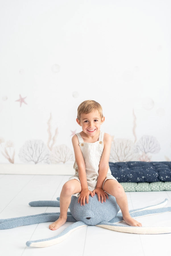 A young boy smiling, seated on a large, plush Octopus Stuffed Animal in a children's room decorated with a sea theme, including starfish and corals on the wall.