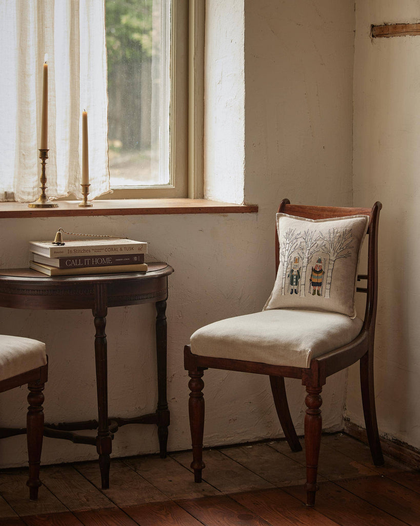 A cozy corner features a wooden chair with the Coral & Tusk Aspen Skiers Pillow, a small table with books and tall candles, all bathed in natural light from a window with sheer curtains—perfect for winter decor.