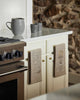 In a kitchen, the Coral & Tusk Snowshoe Hare Tea Towel hangs on a cabinet handle beside another tea towel, with a gray pitcher and cups on a white countertop near a stainless steel stove, and a stone wall in the background.