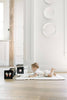 A baby enjoys tummy time on a soft mat placed on a wooden floor, gazing at Baby's Tummy Time Sports black-and-white pattern cubes for visual stimulation. The bright room features white decor and plates displayed on the wall.