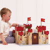 A young child in a white shirt engages in imaginative play with the Lionheart Wooden Castle, crafted from sustainable wood and adorned with red towers, shields, and lion flags, while sitting on the floor against a white background.