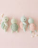 Four heirloom-quality Cuddle + Kind Baby Sea Turtle (Kelp Green) plush toys are arranged in a row on a light pink background, surrounded by small white seashells.
