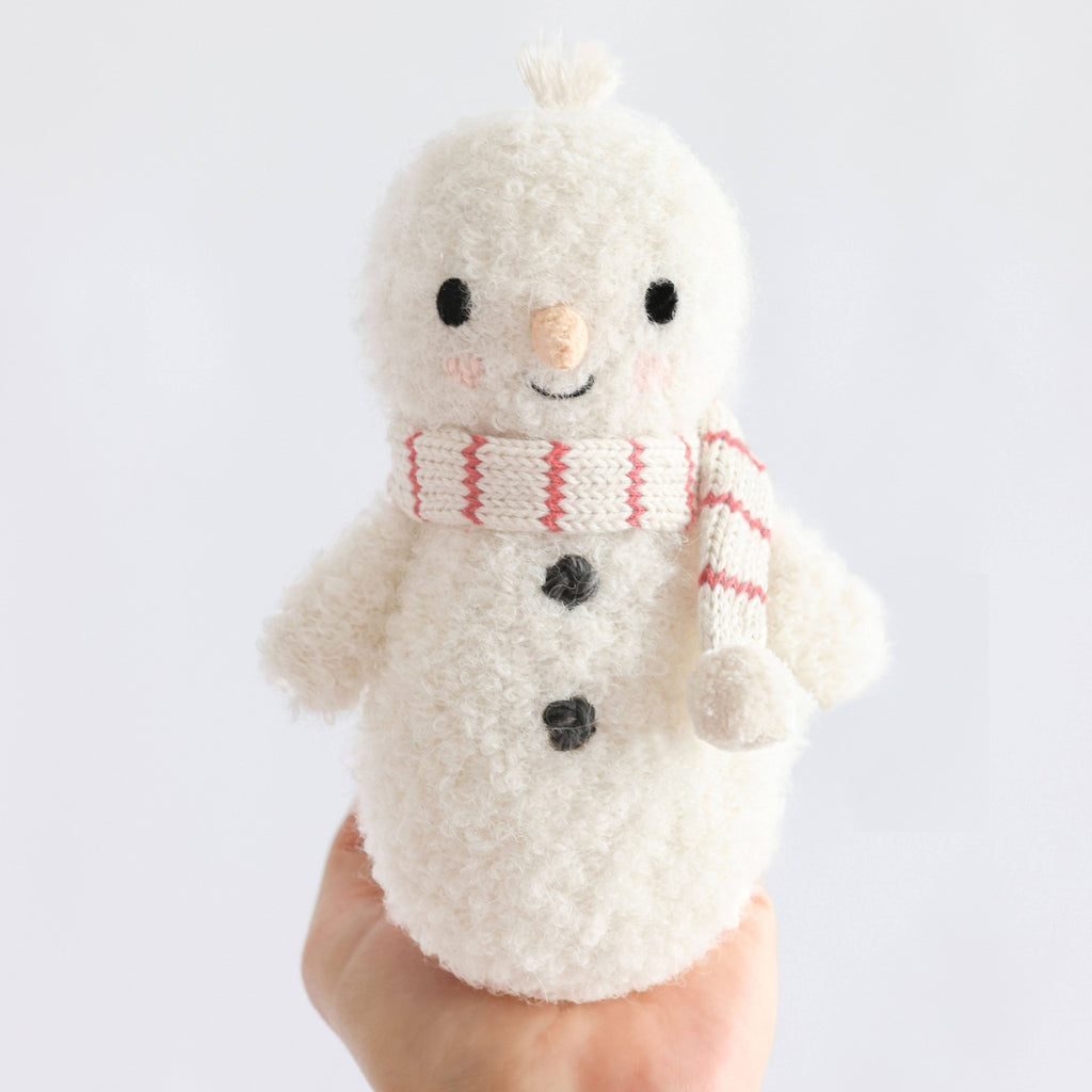 A hand holds the Cuddle + Kind Baby Snowman - Candy Cane, a small plush snowman with a pink and white striped scarf, black button eyes, gray buttons, and an orange nose—set against a plain white background.