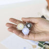 A hand holds three small paint pots labeled 12, 9, and 8—ideal for the Paint By Number Kit - Rome. The background is softly blurred in light hues, sparking creativity for aspiring artists.