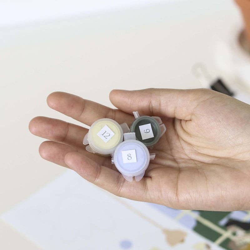 A hand holds three small paint pots labeled 12, 9, and 8—ideal for the Paint By Number Kit - Rome. The background is softly blurred in light hues, sparking creativity for aspiring artists.