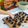 Two children shape dark, round dough balls by hand on parchment-lined baking trays outside. Their messy hands and a green bowl show them enjoying a creative STEM project with the DIY Seed Ball Kit - Rainbow Garden.