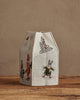 The Coral & Tusk Snow Day Tissue Box Cover is a fabric lantern shaped like a milk carton, embroidered with whimsical skiing rabbit and fox characters. Set on a wooden surface against a beige wall, it’s perfect for snow day decor.