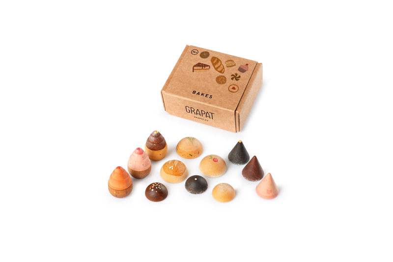 A set of small, colorful, cone-shaped sustainable wooden toys from the Grapat Play Set - Bakes is arranged in front of a brown cardboard box labeled "BAKES GRAPAT" on a white background.