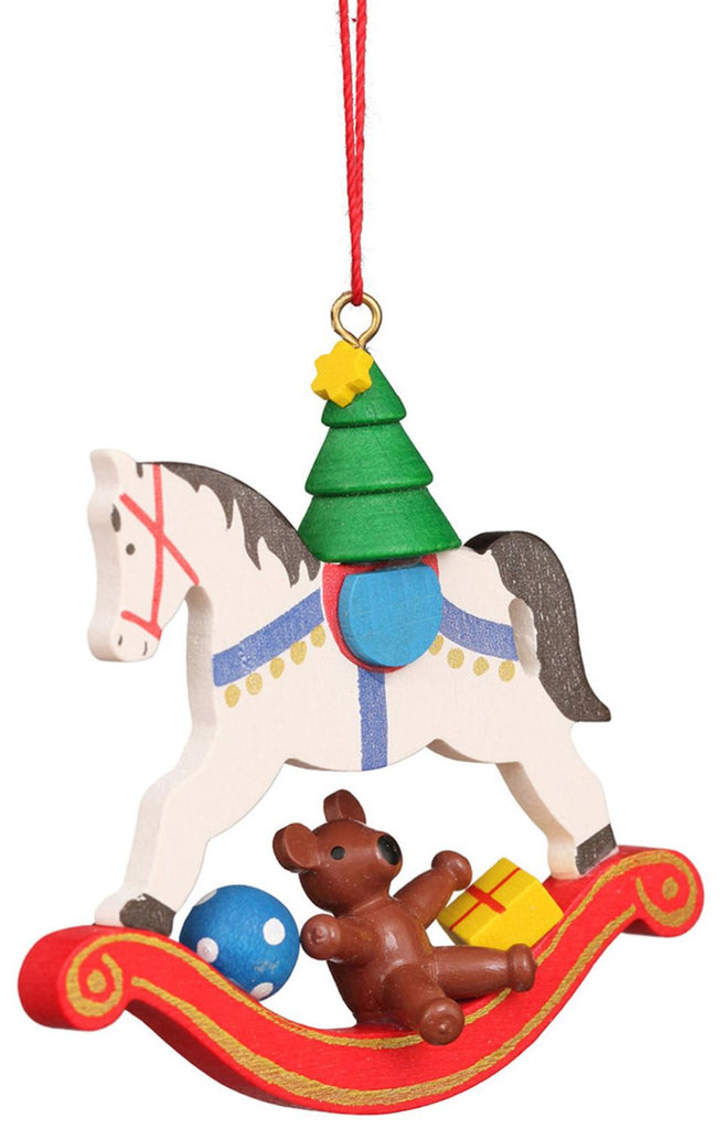 The Christian Ulbricht Ornament - Rocking Horse with Toys features a vibrant hand-painted white wooden rocking horse adorned with a green Christmas tree on its back. A brown teddy bear clutching a yellow gift sits gracefully on the red rocker, joined by a blue ball, while a red string elegantly suspends this delightful decoration.