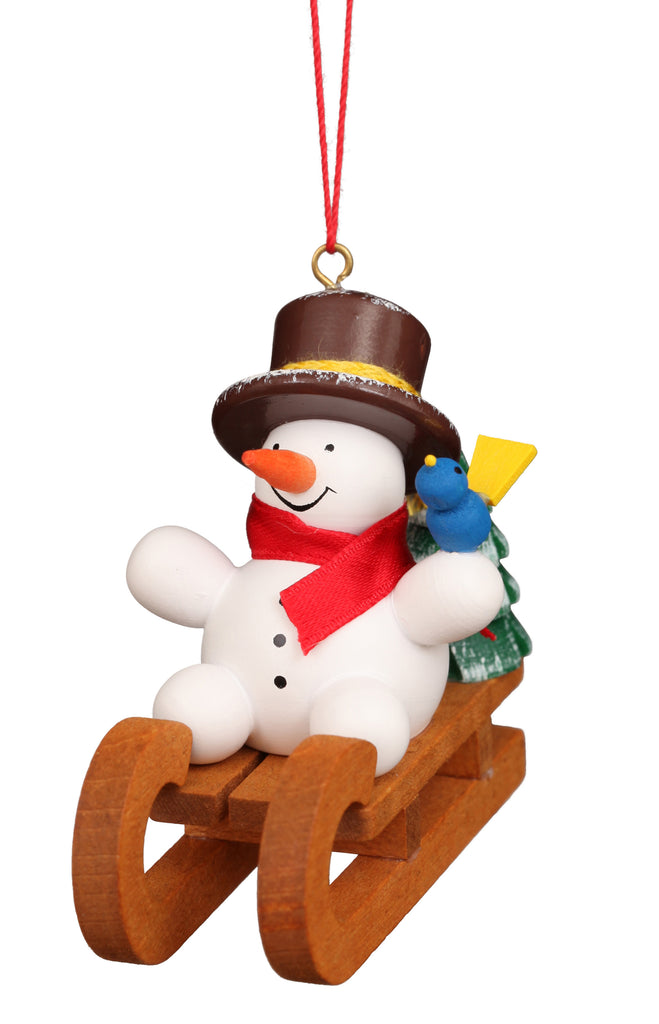 The Christian Ulbricht Ornament - Snowman on Sled features a snowman with a red scarf and brown hat, holding a green tree on a wooden sled, a blue and yellow bird on its arm, and hangs by a red string. Fine German craftsmanship.