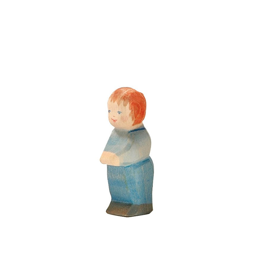 An Ostheimer Toddler figurine of a person with short, red hair. The figurine is dressed in blue pants and a light blue shirt, standing upright with arms folded. Made from sustainably sourced materials and inspired by Ostheimer toys, it features a plain white background.