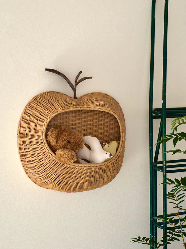 The Ferm Living Apple Braided Wall Pocket - Natural holds a brown teddy bear and a white plush toy—ideal for children's room storage. A green plant and part of a green metal ladder are visible on the right side.