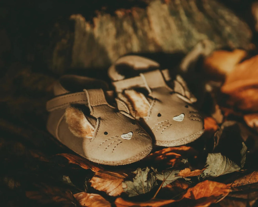 A pair of small, beige Donsje Leather Spark Exclusive Lined Shoes with Squirrel face designs, crafted from premium leather, lying among autumn leaves under warm, soft lighting.